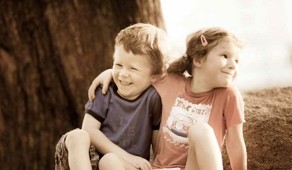 A young boy and girl, about 5 years old, are sitting on a large rock in the sunshine. They are both smiling sheepishly - the girl has her arm around the boy.
