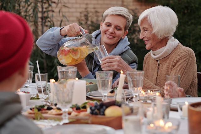 a young woman is sitting at an outdoor dinner table and pouring her grandmother a glass of iced tea. The table is set elegantlyoutddors with white tablecloth and white candles. 