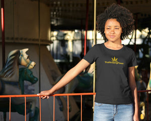 A woman standing next to a ferris wheel looks to her right and smiles. She is wearing a black T-shirt and jeans. The T-shirt has a TheAmazingFlower.com  gold hemp leaf logo on the front..