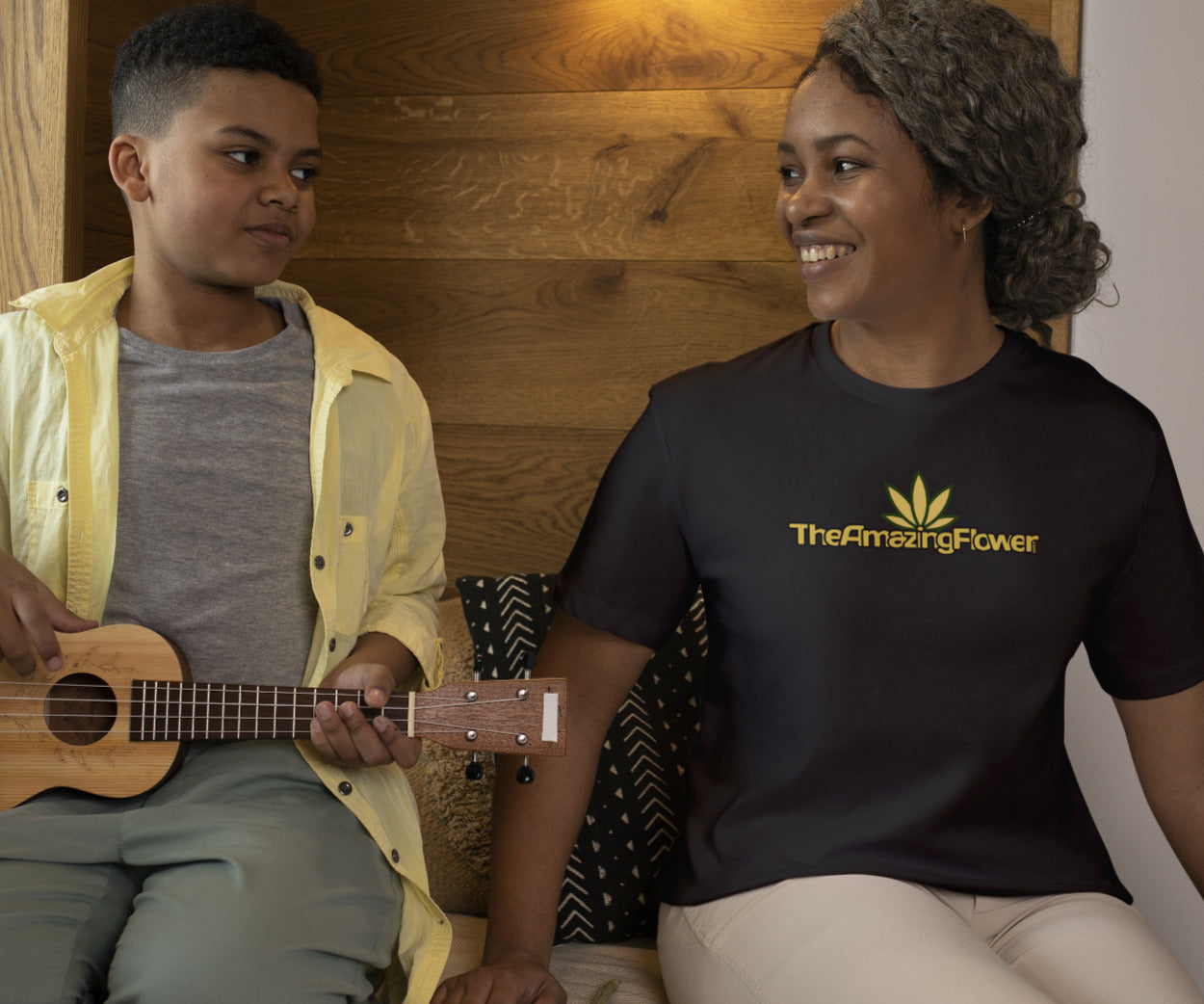 Old Gold Hemp Leaf Logo Women's T-Shirt in black worn by a woman looking at her son playing a ukulele