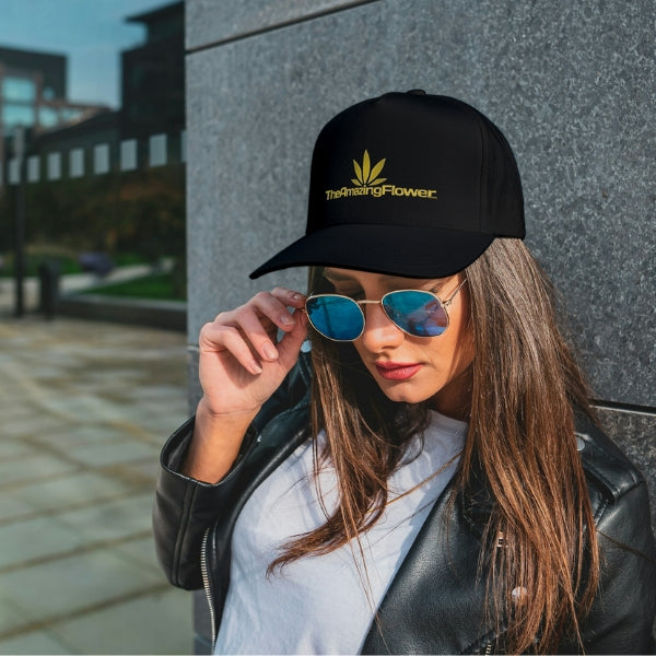 Black baseball cap with gold TheAmazingFlower.com hemp leaf logo worn by a woman wearing a white Tshirt and black leather jacket leaning against a granite wall and looking down over the top of her sunglasses