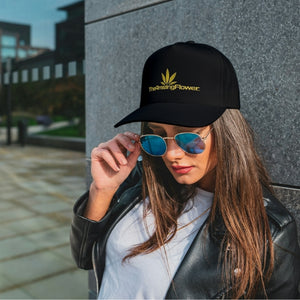 Open image in slideshow, Black baseball cap with gold TheAmazingFlower.com hemp leaf logo worn by a woman wearing a white Tshirt and black leather jacket leaning against a granite wall and looking down over the top of her sunglasses
