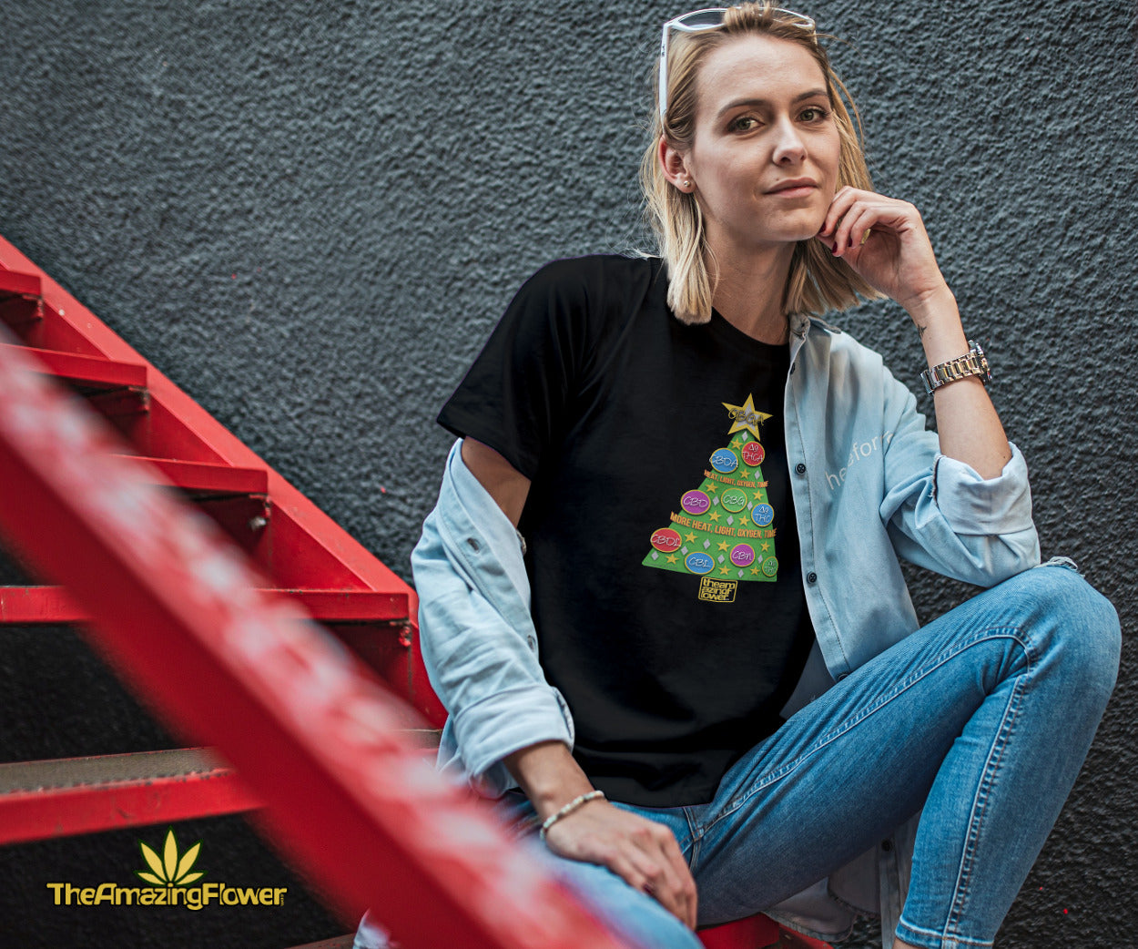 Cannabinoid Holiday Tree Women's Black T-Shirt worn by a blonde woman in jeans sitting on red metal stairs. 